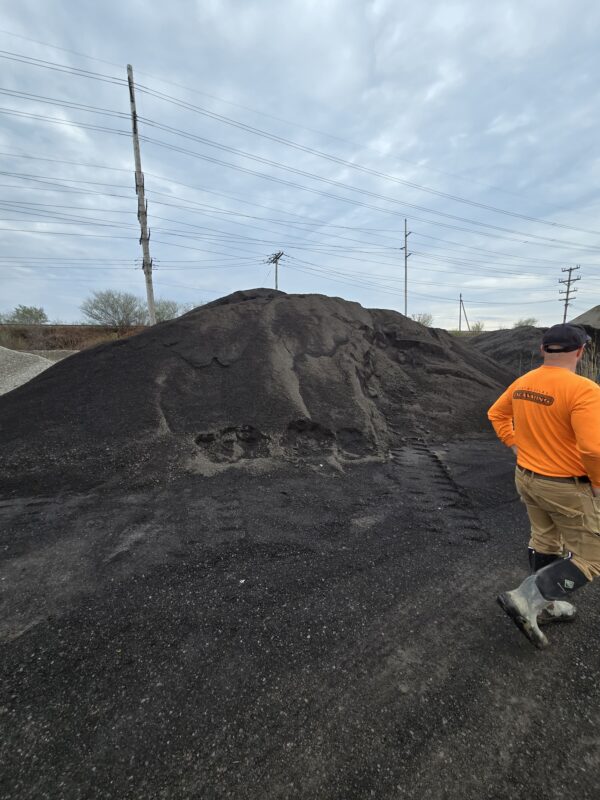 20260416_105415 - wnyDIRT.com Worker in an orange shirt and tan pants stands near a large pile of coal on a gravel lot, with power lines and posts in the background.