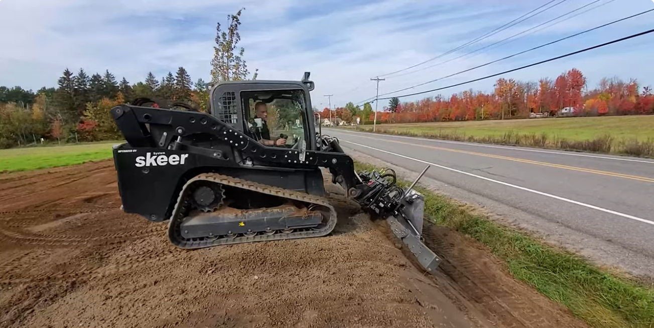 Tracked skid-steer loader (Skeeer) digging a roadside trench beside a two-lane road amid fall-colored trees, operator inside cab overseeing the task.